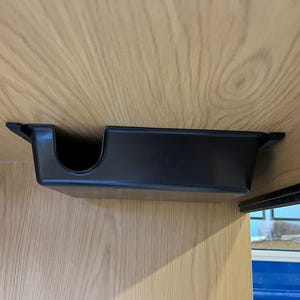 May include: A black plastic cup holder mounted under a wooden surface. The holder has a rectangular shape with a curved opening on one side. The wood has a light brown color and a visible wood grain pattern.