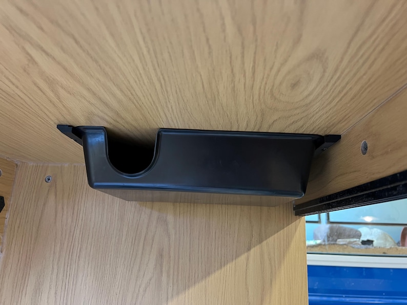 May include: A black plastic cup holder mounted under a wooden surface. The holder has a rectangular shape with a curved opening on one side. The wood has a light brown color and a visible wood grain pattern.