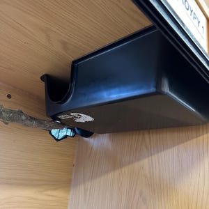 May include: A black plastic reptile enclosure accessory, featuring a curved edge and a rectangular shape. The accessory is mounted inside a wooden enclosure, with a branch extending into the space. The enclosure has a label that reads "ROYPY 1".