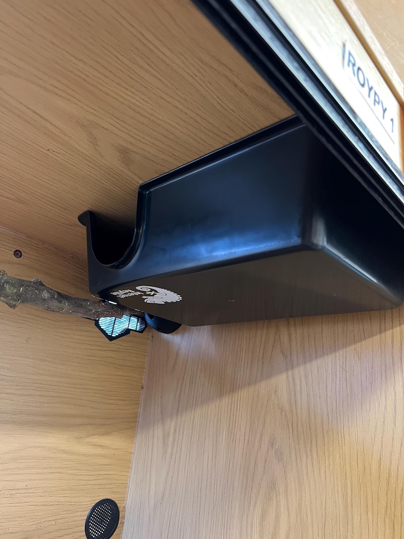 May include: A black plastic reptile enclosure accessory, featuring a curved edge and a rectangular shape. The accessory is mounted inside a wooden enclosure, with a branch extending into the space. The enclosure has a label that reads "ROYPY 1".