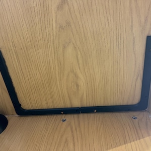 May include: A close-up shot of a wooden shelf with a black metal bracket and a tree branch. The bracket is U-shaped and attached to the shelf with screws. The branch is positioned on the left side of the shelf.