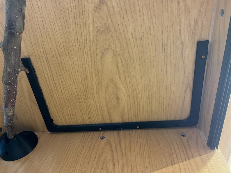 May include: A close-up shot of a wooden shelf with a black metal bracket and a tree branch. The bracket is U-shaped and attached to the shelf with screws. The branch is positioned on the left side of the shelf.