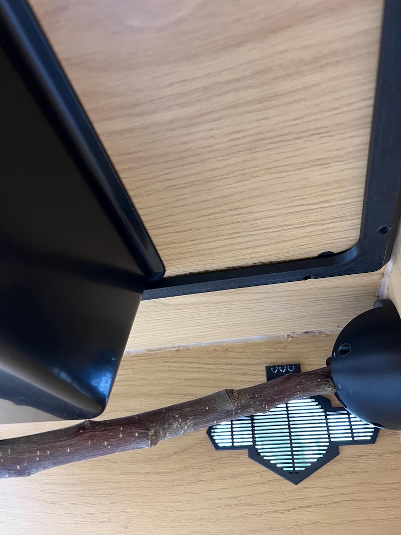 May include: A close-up view of a reptile enclosure interior. The image shows a black plastic corner, a wooden branch, and a black ventilation system. The background is light-colored wood.