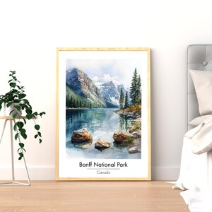 May include: A framed watercolor print of Banff National Park in Canada. The artwork depicts a serene lake surrounded by mountains, trees, and rocks. The text "Banff National Park" and "Canada" are printed at the bottom of the image. The frame is a light wood color.