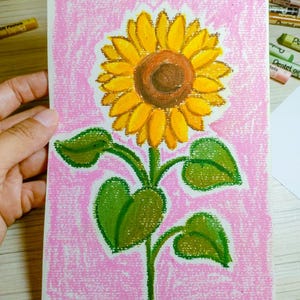 Pode incluir: Ilustração de girassol desenhada à mão sobre fundo rosa. O girassol tem pétalas amarelas, centro castanho e folhas e caule verdes. A obra de arte é criada com pastéis a óleo sobre papel.