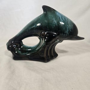 May include: A ceramic dolphin ornament in dark teal and black. The dolphin is shown leaping over a wave, with a glossy finish. The ornament has a smooth, curved design and is likely a decorative item.