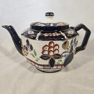 May include: A decorative ceramic teapot featuring a floral design. The teapot has a white, crackled base with brown, blue, and gold floral patterns. The handle and spout are dark blue, and the lid has a small, round knob.