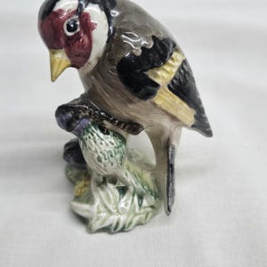 May include: A ceramic goldfinch figurine perched on a branch. The bird features a red face, black and yellow wings, and a yellow beak. The branch is green and purple, and the figurine is set against a white backdrop.