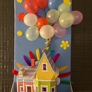 Può includere: Una colorata scena di carta fatta a mano con una casa con tetto giallo e pareti verdi e rosse, adornata con palloncini di vari colori. Fiori e piume decorative aggiungono al design stravagante. I palloncini sono attaccati alla casa.