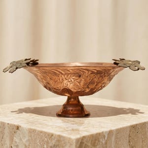 May include: A copper-coloured decorative bowl with a pedestal base and two bird-shaped handles. The bowl has a textured, embossed floral design. The bowl is displayed on a marble surface.