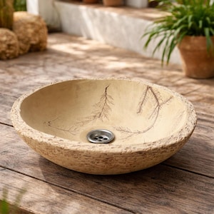 Puede incluir: Un lavabo beige de forma ovalada con una superficie texturizada y un desagüe metálico. El lavabo presenta un diseño decorativo de ramas y se encuentra sobre una superficie de madera. El lavabo es probablemente para uso en exteriores.