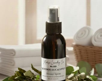 May include: A dark brown glass bottle of Marja Laundry Co. Menthol Eucalyptus Shower Spray, No. 323, with a white label. The bottle is surrounded by fresh green leaves, white towels, and smooth stones, suggesting a spa-like atmosphere.