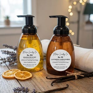 May include: Two clear, amber-colored foaming hand soap dispensers with black pumps. One is labeled "Lemon Delight," the other "Lavender Sandalwood." Both have white labels with black text. Dried orange slices and vanilla beans are arranged nearby.