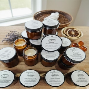 May include: A collection of small, brown glass jars filled with organic deodorant cream. The jars have black lids and white labels with product information. Some jars are open, revealing the cream inside. The image includes dried orange slices and lavender sprigs.