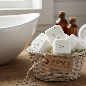 Könnte beinhalten: Ein Weidenkorb gefüllt mit weißen, quadratischen Toilettenbomben, übersät mit Lavendel. Ein braunes Etikett lautet "MARSULAUNDRYCO. Homemade Toilet Bombs." Der Korb steht auf einer Holzoberfläche, in der Nähe eines weißen Waschbeckens und brauner Flaschen.