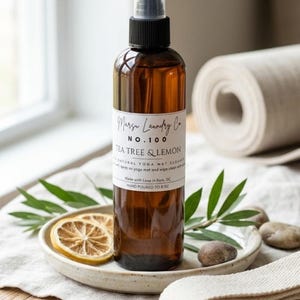 May include: A brown glass spray bottle of yoga mat cleaner with a black cap, labeled "Tea Tree & Lemon." The bottle sits on a small plate with lemon slices and greenery. A rolled yoga mat is in the background.