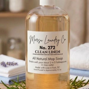 Pode incluir: Uma garrafa transparente de sabão para esfregona natural Clean Linen da Marsu Laundry Co. O rótulo diz "No. 272" e "Shake well, pour about 2 to 3 Tablespoons in mop water." A garrafa tem aproximadamente 946 ml e é rodeada por lavanda e toalhas dobradas.