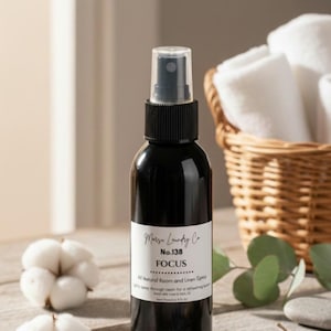 May include: A black room and linen spray bottle with a white label that reads "Focus". The bottle is on a wooden surface with cotton buds, a basket of rolled white towels, and sprigs of lavender and eucalyptus.