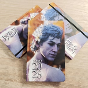 May include: Three 2026 agenda notebooks with a portrait of a person with blue hair and a flower. The notebooks have a black elastic band and the text "BONNIE'S AGENDA" and "20 26". The background is a light wood grain.