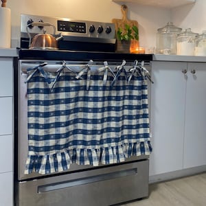 May include: A blue and white gingham kitchen curtain with a frilled edge is tied to an oven. The curtain is made of fabric and has blue ties. A copper kettle sits on the hob.