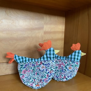 Peut inclure: Deux maniques en forme de poulet. Le haut des poulets est à carreaux verts et blancs, avec des plumes de queue et des crêtes orange. Le bas est un motif floral avec des fleurs roses, bleues et blanches.