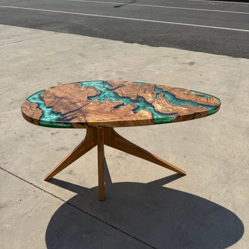May include: Oval wooden coffee table with a unique design. The table features a natural wood grain top with a river of teal-colored epoxy resin. The table is supported by three wooden legs.