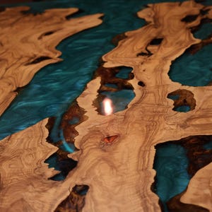 May include: Close-up of a wooden table with a teal epoxy resin river design. The wood has a natural grain pattern with varying shades of brown and tan. The resin creates a contrasting, glossy, and reflective surface.