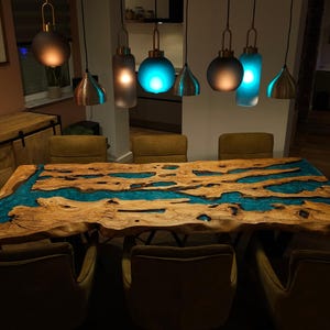 May include: A handcrafted wooden dining table with a river of turquoise resin. The live edge table is surrounded by six olive green chairs. Above the table hang six pendant lights with copper and frosted glass shades.