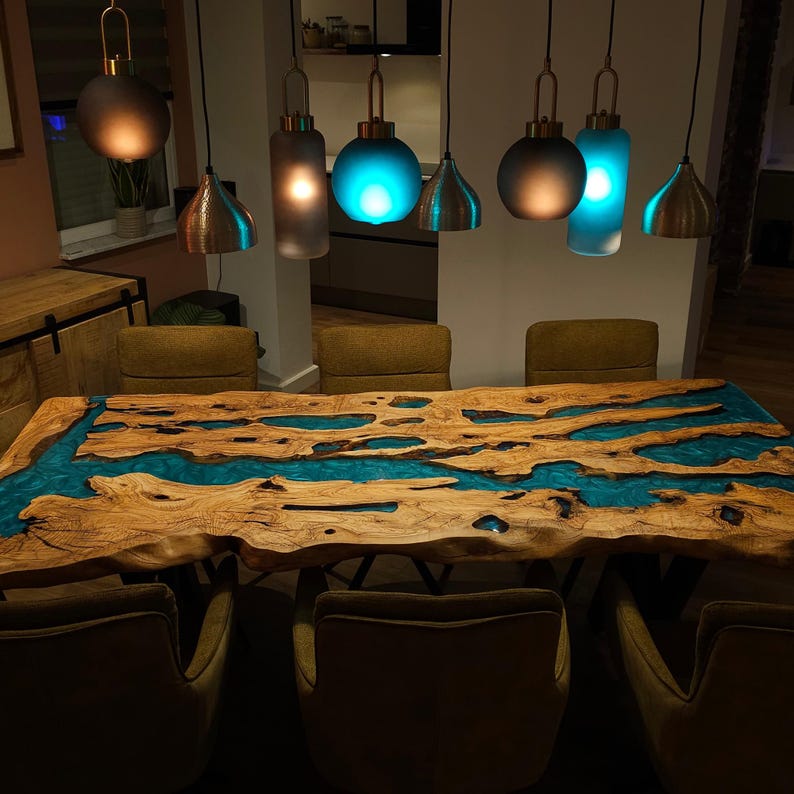 May include: A handcrafted wooden dining table with a river of turquoise resin. The live edge table is surrounded by six olive green chairs. Above the table hang six pendant lights with copper and frosted glass shades.