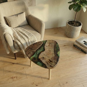 May include: A unique, triangular coffee table with a wood grain top and green resin accents. The table has three light-colored wooden legs. The table is next to a beige armchair with a throw blanket and pillow.