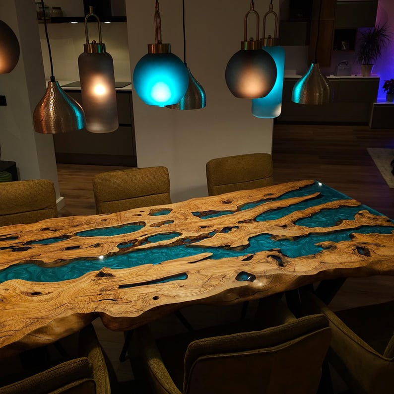 May include: A large, live-edge wooden dining table with a river of turquoise resin flowing through the center. The table is surrounded by brown chairs and illuminated by hanging pendant lights with copper and frosted glass shades.