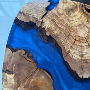 May include: Close-up of a wooden table with a river of blue resin. The wood has a natural grain pattern with shades of brown and tan. The resin flows through the center, creating a striking contrast.