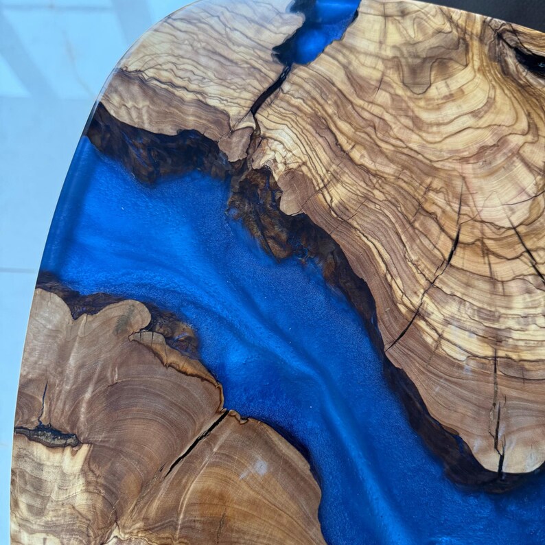 May include: Close-up of a wooden table with a river of blue resin. The wood has a natural grain pattern with shades of brown and tan. The resin flows through the center, creating a striking contrast.