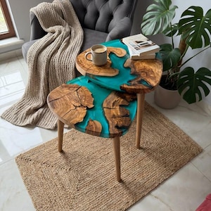 May include: A set of two wooden side tables with a unique design. The tabletops feature a turquoise resin river effect, surrounded by natural wood grain. The tables have light-colored wooden legs and are placed on a woven jute rug.