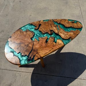 May include: Oval-shaped wooden coffee table with a unique design. The table features a combination of natural wood and teal-colored resin. The wood has a rich, brown color with intricate grain patterns. The table is supported by four wooden legs.