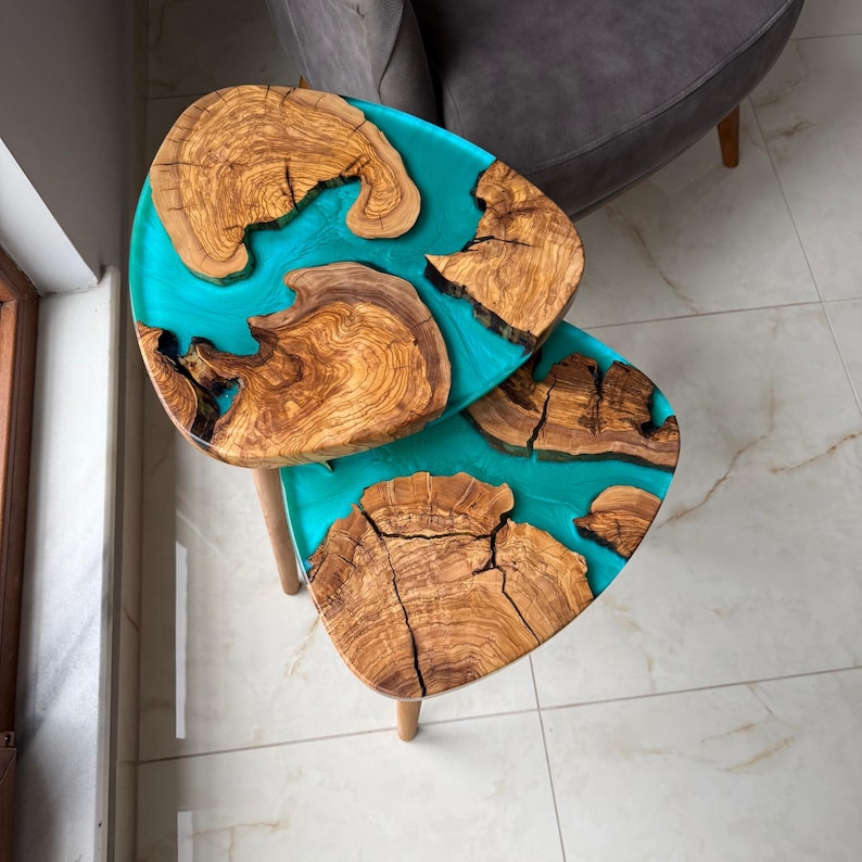 May include: Two wooden side tables with a turquoise resin inlay. The tables have a unique organic shape, with sections of natural wood grain set within the vibrant turquoise resin. The tables are supported by three wooden legs.