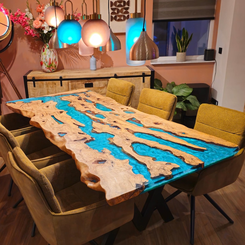 May include: A large, live-edge dining table with a river of turquoise resin. The table is made of light-colored wood with a natural edge, supported by black metal legs. The table is surrounded by mustard-colored chairs.