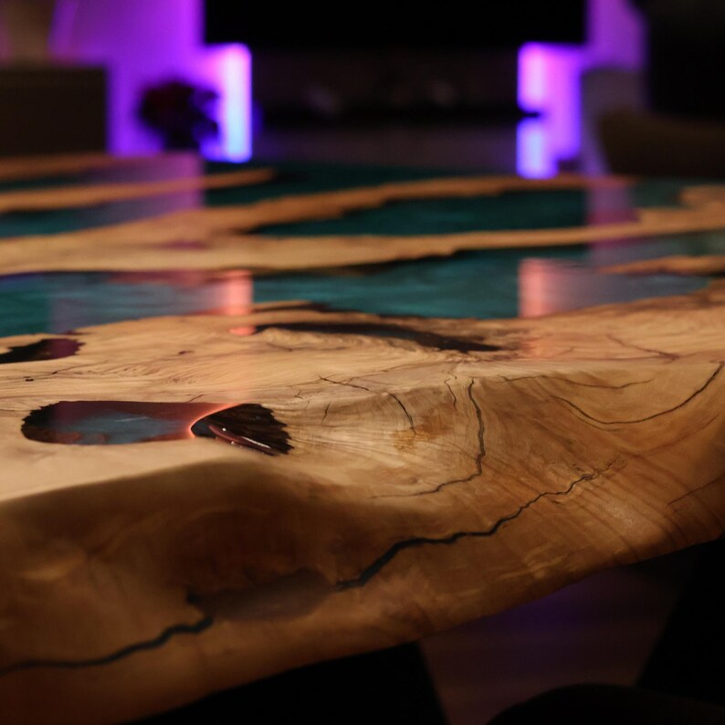 May include: Close-up of a wooden table with a live edge design. The table features a teal-colored epoxy resin river that runs through the center. The wood has a natural, light brown color with visible grain and cracks.