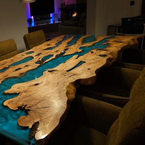 May include: A large, custom-made wooden table with a unique design. The table features a natural wood grain surface with a river of blue epoxy resin flowing through the center. The table is surrounded by chairs.