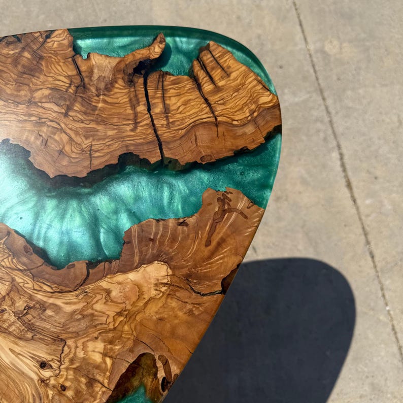 May include: Close-up of a wooden table with a unique design. The table features a combination of natural wood grain and a vibrant teal-colored epoxy resin. The wood has a warm, brown tone, and the resin creates a river-like effect. The table's dimensions are not visible.