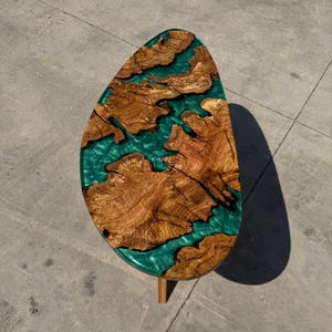 May include: An oval-shaped coffee table with a wooden top and teal-colored resin filling the gaps. The wood has a natural grain pattern. The table is supported by a single wooden leg. The table measures approximately 48 inches long and 24 inches wide.