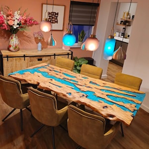 May include: A live edge wooden dining table with a vibrant blue epoxy river running through the center. The table is surrounded by six mustard-colored chairs. Overhead, several pendant lights illuminate the room, creating a warm and inviting atmosphere.