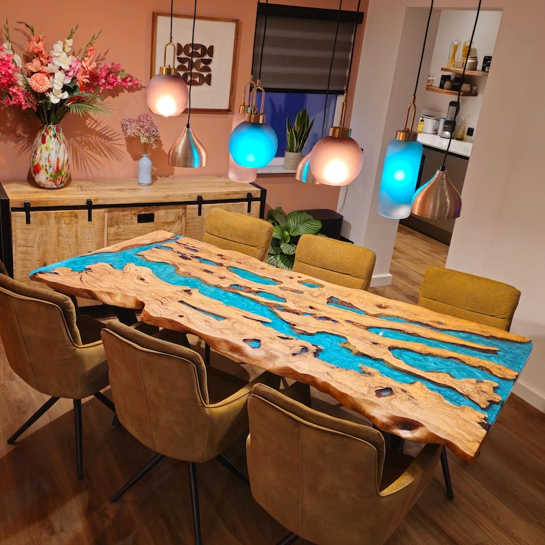 May include: A live edge wooden dining table with a vibrant blue epoxy river running through the center. The table is surrounded by six mustard-colored chairs. Overhead, several pendant lights illuminate the room, creating a warm and inviting atmosphere.