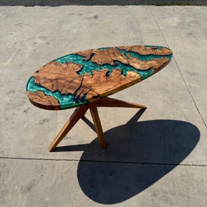 May include: Oval-shaped wooden coffee table with a teal epoxy resin river design. The table's surface is a combination of natural wood grain and a glossy teal resin. The table is supported by three wooden legs.