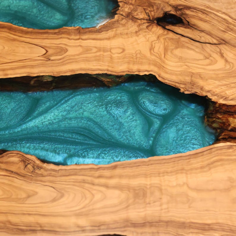 May include: Close-up of a wooden table with a turquoise epoxy resin river. The wood has a natural grain and a light brown color. The resin has a glossy finish and a swirling pattern.
