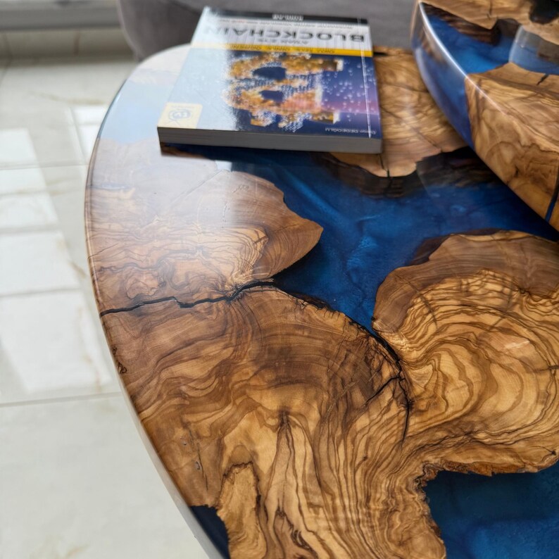 May include: Close-up of a wooden table with a blue resin river design. The table has a natural wood grain pattern with a glossy finish. A book titled "BLOCKCHAIN" rests on the table's surface.
