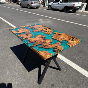 May include: A rectangular wooden table with a turquoise epoxy resin center and natural wood grain patterns. The table is supported by black metal legs. The wood has a warm brown color, contrasting with the vibrant blue-green resin.