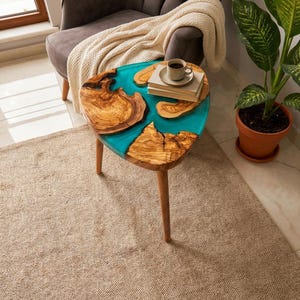 Puede incluir: Una mesa auxiliar triangular única con una tapa de madera y una incrustación de resina turquesa. La mesa presenta patrones de vetas de madera natural y tres patas de madera. Una taza de café y libros descansan sobre la mesa. La mesa mide unos 61 cm de ancho.