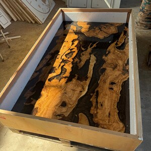 May include: A large wooden table top with a unique design. The table top features a combination of light brown wood and dark black resin. The wood has a natural grain pattern, and the resin fills in the gaps. The table top is inside a wooden frame.