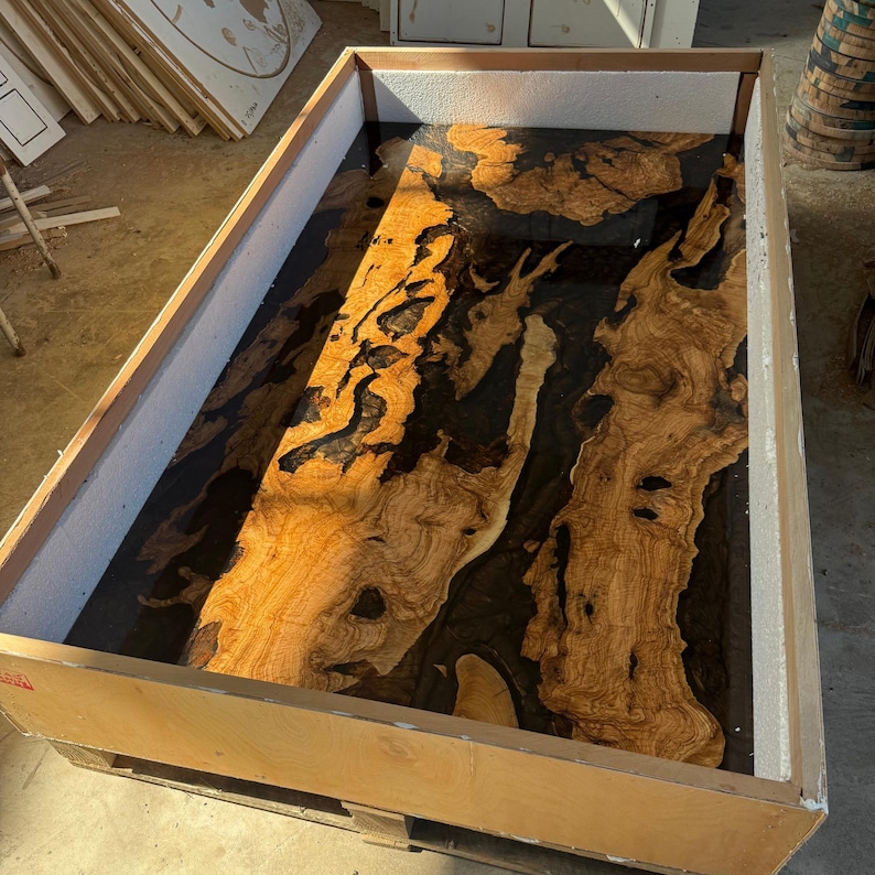 May include: A large wooden table top with a unique design. The table top features a combination of light brown wood and dark black resin. The wood has a natural grain pattern, and the resin fills in the gaps. The table top is inside a wooden frame.